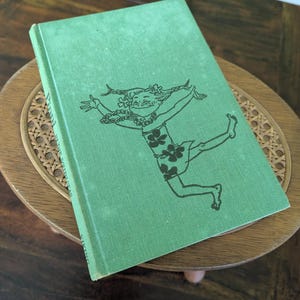 Pu&ograve; includere: Un libro verde vintage con un'illustrazione nera di una figura danzante sulla copertina. Il libro &egrave; appoggiato su un tavolino rotondo in legno con un piano intrecciato. Il titolo del libro &egrave; parzialmente visibile.