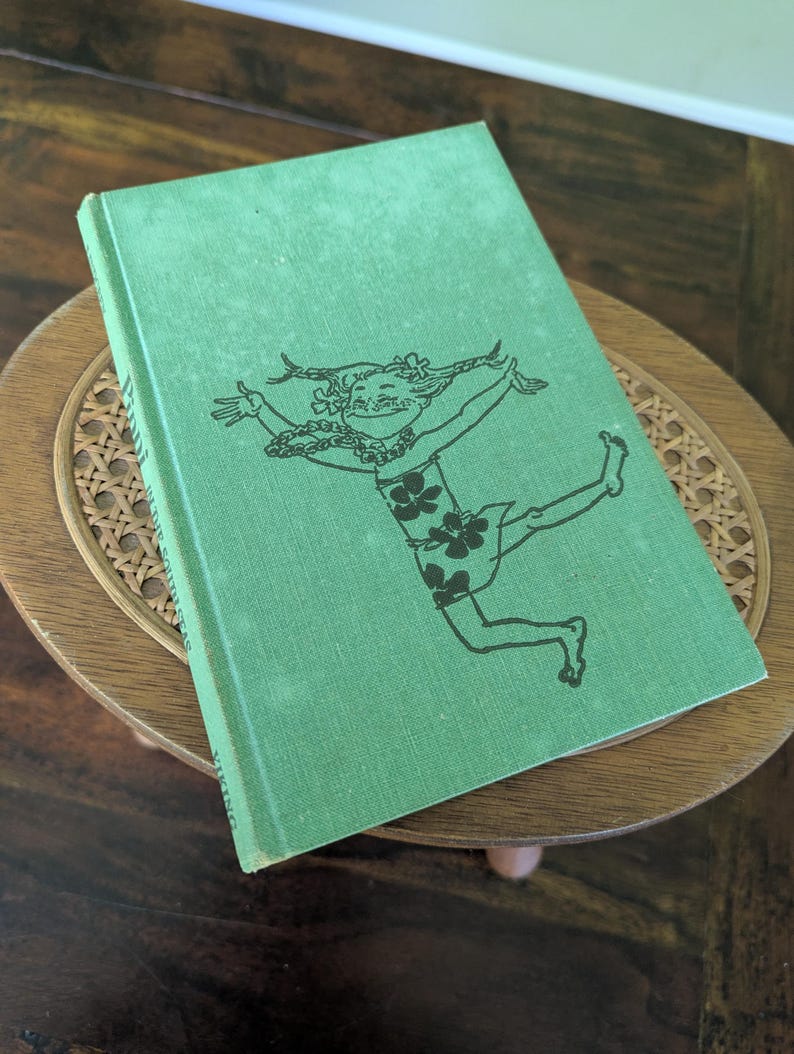 Pu&ograve; includere: Un libro verde vintage con un'illustrazione nera di una figura danzante sulla copertina. Il libro &egrave; appoggiato su un tavolino rotondo in legno con un piano intrecciato. Il titolo del libro &egrave; parzialmente visibile.