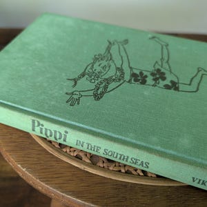 Può includere: Un libro vintage con copertina rigida intitolato "Pippi in the South Seas" di Lindgren. Il libro ha una copertina verde con un'illustrazione nera di un personaggio. Il libro è appoggiato su una superficie di legno.