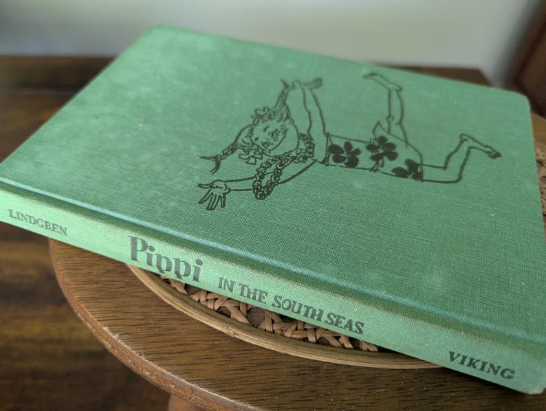 Pu&ograve; includere: Un libro vintage con copertina rigida intitolato "Pippi in the South Seas" di Lindgren. Il libro ha una copertina verde con un'illustrazione nera di un personaggio. Il libro &egrave; appoggiato su una superficie di legno.