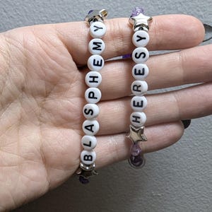 May include: Two white beaded bracelets with silver star charms. The bracelets spell out the words "blasphemy" and "heresy".