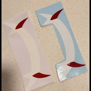 May include: Two white vinyl decals with red accents. The decals are shaped like a curved arrow with a pointed tip. The decals are on a light blue and white background.