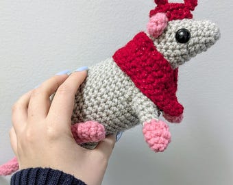 May include: A crocheted rat toy in gray, red, and pink. The rat has a red scarf and hat, pink feet and ears, and a pink tail. The toy is held in a hand against a white background.