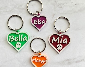 May include: Four heart-shaped dog tags with white paw prints and names "Elsa", "Bella", "Mia", and "Mango" printed in white. The tags are attached to silver key rings.