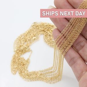 May include: A close-up of a gold chain necklace. The chain is made of small, delicate links and is shown in a hand, with multiple strands of the chain laid out on a white surface. The text "SHIPS NEXT DAY" is visible in the top right corner of the image.