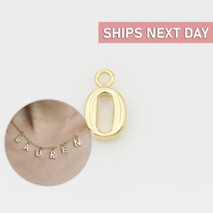 May include: Gold-coloured metal charm in the shape of the number zero. The charm has a slightly rounded, oval shape and is designed to be added to a necklace or bracelet.