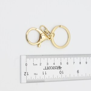 May include: Gold-toned metal key chain with a swivel clasp and a round ring. The key chain is measured against a ruler.