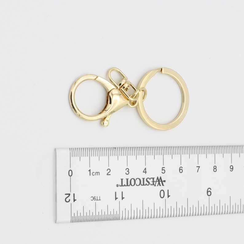 May include: Gold-toned metal key chain with a swivel clasp and a round ring. The key chain is measured against a ruler.