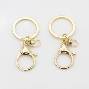 May include: Two gold-toned keychains with a round ring and a lobster clasp. The keychains are made of metal and have a shiny finish.