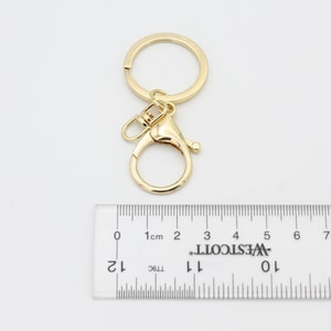 May include: Gold-toned key chain with a large, round ring and a smaller, round swivel clasp. The key chain is measured against a ruler with markings in centimeters.