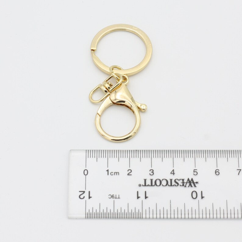 May include: Gold-toned key chain with a large, round ring and a smaller, round swivel clasp. The key chain is measured against a ruler with markings in centimeters.