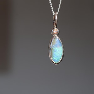 Puede incluir: Un collar con colgante de plata que presenta un ópalo ovalado con destellos iridiscentes azules y verdes. El ópalo está engastado en un bisel de plata liso, rematado con una pequeña piedra preciosa redonda de color marrón claro, en una cadena.