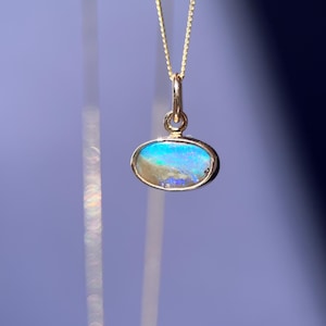Könnte beinhalten: Eine goldene Halskette mit Anhänger, die einen ovalen Opal-Edelstein trägt. Der Opal zeigt schillernde Farben von Blau, Grün und Gold. Der Anhänger hängt an einer zarten Goldkette und bildet ein elegantes Schmuckstück.