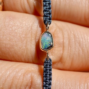 Pode incluir: Uma pulseira com uma opala central de formato irregular engastada num bisel de metal dourado. A pedra apresenta reflexos iridescentes em tons de verde e azul. A pulseira é feita de um cordão entrançado de cor azul-escura.