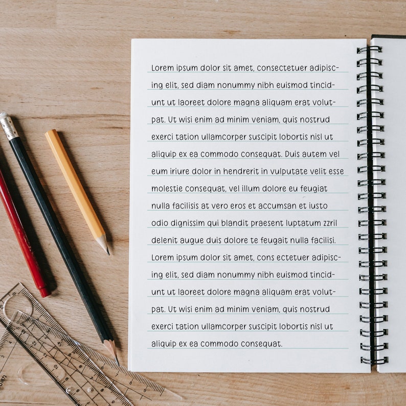 Puede incluir: Un cuaderno espiral abierto, con p&aacute;ginas rayadas, lleno del texto "Lorem ipsum dolor sit amet...", est&aacute; junto a tres l&aacute;pices y una regla transparente sobre una superficie de madera. Los l&aacute;pices son rojo, negro y amarillo.
