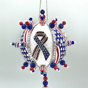 Op de afbeelding: Een witte, rood-blauwe, parelversierde kerstbal met een rood-wit-blauw lint en sterren. De bal heeft een lange parelketting.