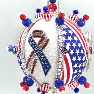 Op de afbeelding: Een witte, ronde kerstbal versierd met rode, witte en blauwe kralen, strass steentjes en een Amerikaans vlag lint. De kerstbal is een patriottische kerstversiering.