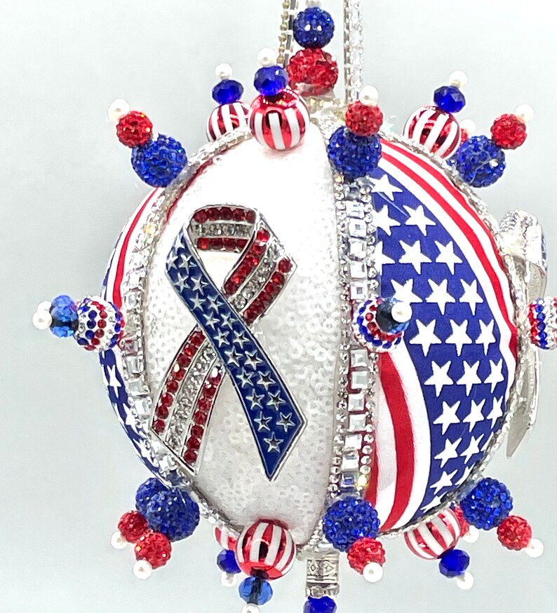 Op de afbeelding: Een witte, ronde kerstbal versierd met rode, witte en blauwe kralen, strass steentjes en een Amerikaans vlag lint. De kerstbal is een patriottische kerstversiering.