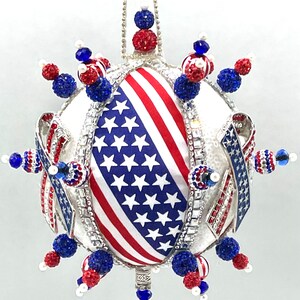 Op de afbeelding: Een witte, ronde kerstbal met een rood, wit en blauw Amerikaans vlagontwerp. De bal is versierd met blauwe en rode kralen en strass.