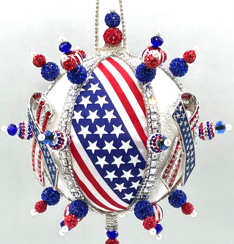 Op de afbeelding: Een witte, ronde kerstbal met een rood, wit en blauw Amerikaans vlagontwerp. De bal is versierd met blauwe en rode kralen en strass.