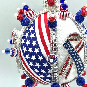 Op de afbeelding: Een witte, ronde, glinsterende kerstbal met een rood-wit-blauw Amerikaans vlagontwerp en een blauw-wit lint met sterren. De kerstbal is versierd met rode, witte en blauwe kralen.