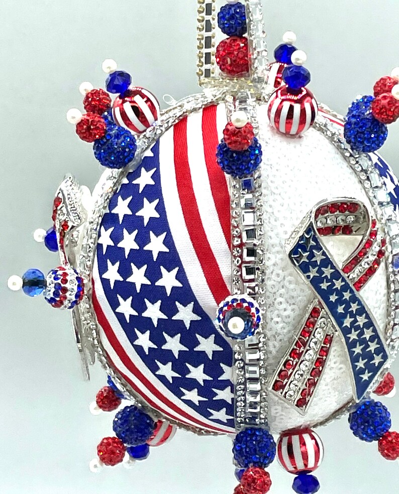 Op de afbeelding: Een witte, ronde, glinsterende kerstbal met een rood-wit-blauw Amerikaans vlagontwerp en een blauw-wit lint met sterren. De kerstbal is versierd met rode, witte en blauwe kralen.