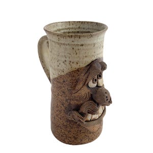 May include: A tall, handmade ceramic mug with a whimsical face sculpted on the front. The mug features a speckled beige and brown colour scheme, with a large handle. The face has exaggerated features, including large eyes and a grinning mouth.