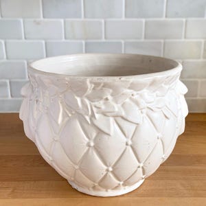 May include: Cream-colored ceramic planter with a quilted diamond pattern and leaf-like accents around the rim. The planter has a wide opening and sits on a wooden surface, ideal for indoor or outdoor plants.