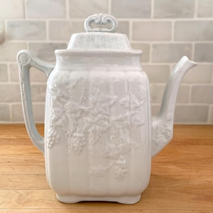 May include: White ceramic teapot with a lid and handle. The teapot has a raised grape vine design on the body and a decorative handle. The spout is curved and the lid has a decorative finial. The teapot is on a wooden surface.