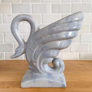 Peut inclure: Un vase en céramique bleu clair en forme de cygne. Le vase a un cou incurvé et un motif en forme d'aile d'un côté. La base est carrée. Le vase est sur une surface en bois avec un fond en briques blanches.