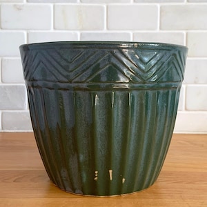 May include: A dark green ceramic planter with a glossy finish. The planter features vertical ribbed detailing and a geometric chevron pattern around the rim. The planter is designed for indoor or outdoor use.