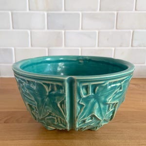 May include: A teal green ceramic planter with a leaf design. The planter is round and has a wide base.