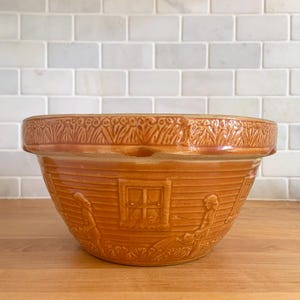 May include: A large, brown ceramic mixing bowl with a raised design of a house with windows and figures. The bowl has a scalloped rim and a textured surface.