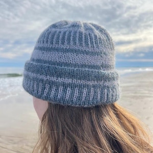 May include: A knitted beanie featuring a ribbed cuff and alternating horizontal stripes in shades of slate grey and light grey. The hat has a textured, fuzzy appearance with a structured crown and a folded brim that sits snugly around the head.