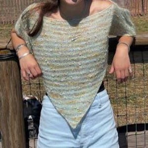 以下が含まれることがあります： ライトグリーンとイエローのニットオフショルダーのトップス。ユニークな三角形の形をしています。トップスは半袖で、テクスチャード加工が施されています。モデルはライトブルーのデニムショーツを着用しています。