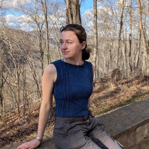 May include: A sleeveless, navy blue knit top with ribbed detailing. The top has a crew neck and a fitted silhouette. The person is wearing gray cargo pants and a silver bracelet. The background features a forest with bare trees and a blue sky.
