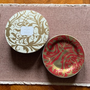 May include: A set of jewel plates with a red and gold design. The plate is round with a gold rim. The box is round with a cream background and gold floral design. The text on the box reads "jewel plates".