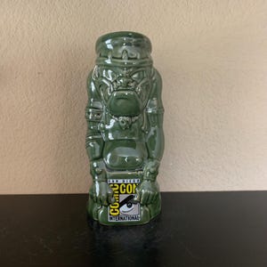 Puede incluir: Taza tiki de cerámica verde que representa una figura estilizada con una expresión severa. La taza tiene un diseño detallado con rasgos faciales y contornos corporales visibles. La base de la taza muestra el logotipo de San Diego Comic-Con International.