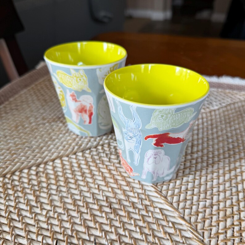 Puede incluir: Dos tazas de cer&aacute;mica con exterior azul claro e interior amarillo brillante. Las tazas est&aacute;n decoradas con varias ilustraciones de perros en rojo, amarillo y blanco. Las tazas est&aacute;n sobre una estera tejida.