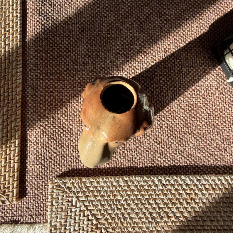 Peut inclure: Un vase en c&eacute;ramique marron avec un design abstrait unique. Le vase a une grande ouverture en haut et une surface textur&eacute;e. Le vase est pos&eacute; sur une surface tiss&eacute;e.