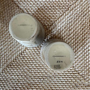 May include: Two cylindrical ceramic mugs with a textured, speckled finish in shades of beige and gray. The bottom of each mug is stamped with the text "ANTHROPOLOGIE". The mugs are resting on a woven, light brown surface.