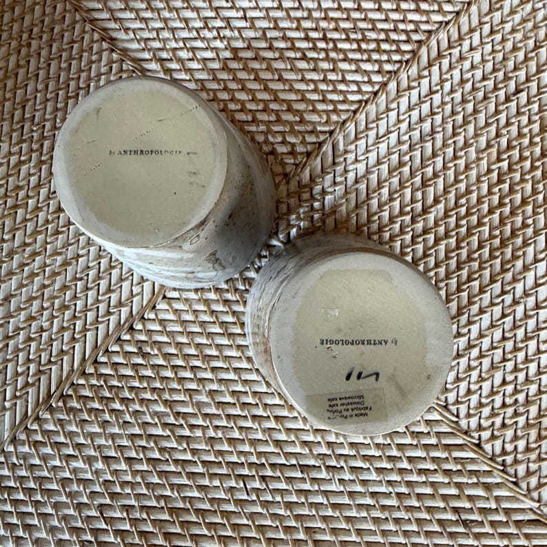May include: Two cylindrical ceramic mugs with a textured, speckled finish in shades of beige and gray. The bottom of each mug is stamped with the text "ANTHROPOLOGIE". The mugs are resting on a woven, light brown surface.