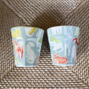 Puede incluir: Dos vasos de cer&aacute;mica azul claro con un patr&oacute;n repetido de ilustraciones de perros coloridos. Los perros est&aacute;n representados en varias poses y colores, incluyendo rojo, amarillo, verde y azul. Los vasos tienen forma hexagonal.