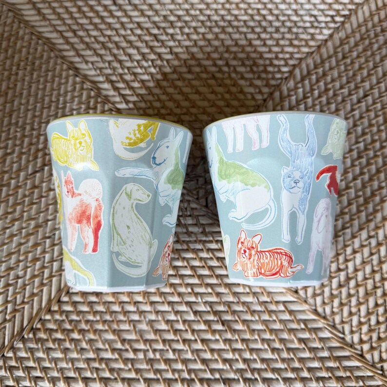 Puede incluir: Dos vasos de cer&aacute;mica azul claro con un patr&oacute;n repetido de ilustraciones de perros coloridos. Los perros est&aacute;n representados en varias poses y colores, incluyendo rojo, amarillo, verde y azul. Los vasos tienen forma hexagonal.