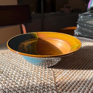 May include: A ceramic bowl with a textured, light blue exterior and a vibrant interior. The interior transitions from a dark teal to a warm orange-yellow hue. The bowl sits on a woven mat.