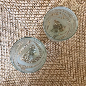 May include: Two clear glass goblets with frosted, swirling designs. The goblets are set on a woven, light brown surface. The design on the glass is a series of curved lines that create a spiral effect.