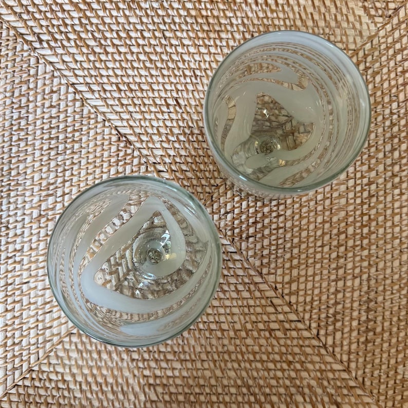 May include: Two clear glass goblets with frosted, swirling designs. The goblets are set on a woven, light brown surface. The design on the glass is a series of curved lines that create a spiral effect.