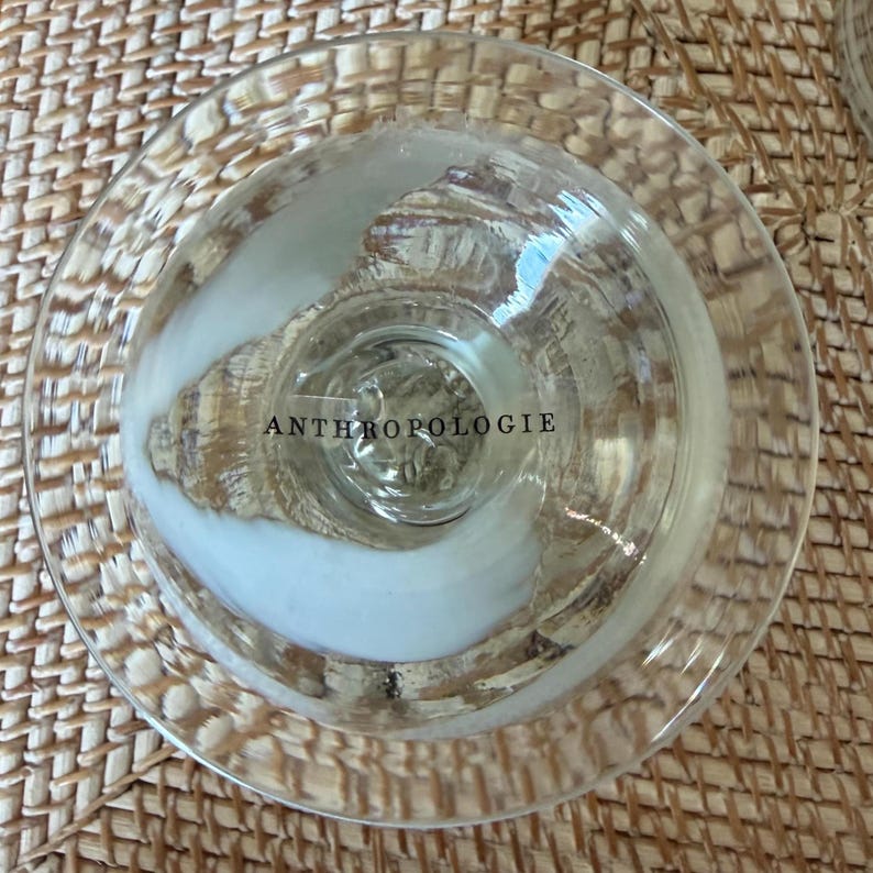 May include: Clear glass goblet with a wide, shallow bowl and a short stem. The word "ANTHROPOLOGIE" is printed on the side of the glass. The goblet is sitting on a woven surface.