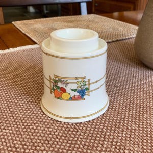 May include: A white ceramic egg cup with a decorative fruit and bamboo design. The egg cup has a cylindrical base and a circular top, with gold trim. The fruit design includes oranges, lemons, and grapes.