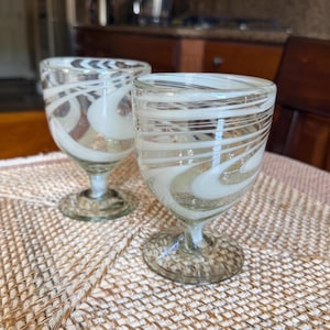 May include: Two clear glass goblets with white swirl designs. The goblets have a rounded bowl shape, a stem, and a circular base. The white swirls create a decorative pattern on the glass. The goblets are sitting on a woven surface.